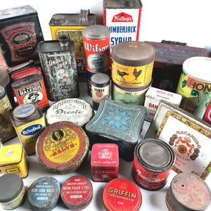 Pode incluir: Uma coleção de latas e recipientes de metal vintage, com várias formas, tamanhos e cores. Algumas latas exibem textos como "MUSTARD", "HONEY" e "BAND-AID". A variedade inclui recipientes redondos, quadrados e retangulares, mostrando designs antigos.