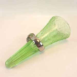 May include: A light green glass wall pocket vase featuring a flared opening with embossed floral patterns and vertical ribbing along the tapered base. A polished silver-toned metal mounting bracket is secured around the center of the glass body.