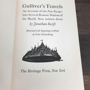 K&ouml;nnte beinhalten: Offenes Buch mit dem Titel "Gulliver's Travels" und Text von Jonathan Swift. Das Buch zeigt eine Illustration einer Stadt und wird von The Heritage Press, New York, herausgegeben.