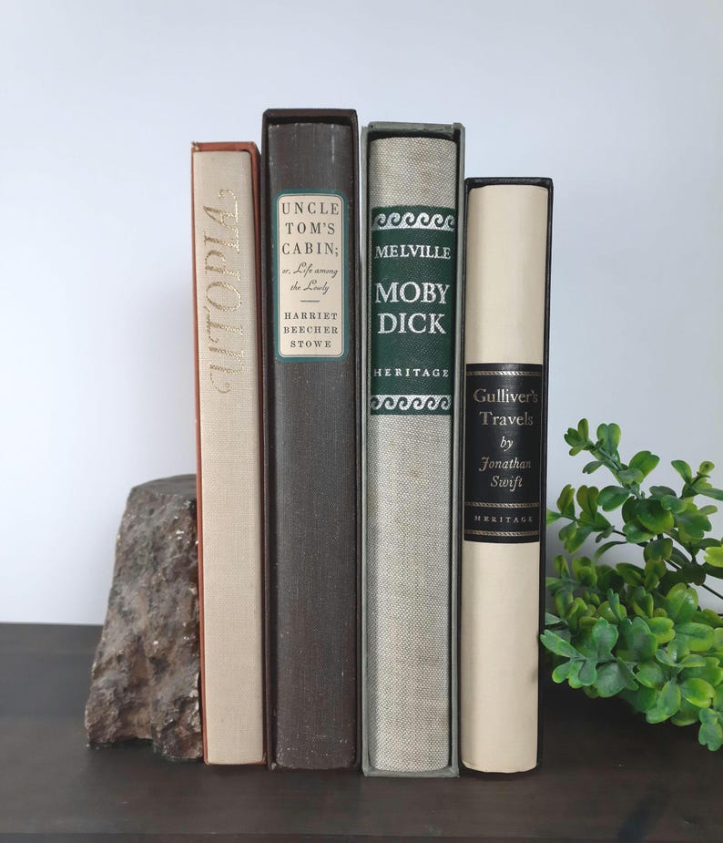 K&ouml;nnte beinhalten: Eine Sammlung alter B&uuml;cher mit Titeln wie "Utopia", "Uncle Tom's Cabin" und "Moby Dick". Die B&uuml;cher sind in verschiedenen Farben wie Beige, Braun und Gr&uuml;n gehalten und vertikal angeordnet. Links befindet sich ein dekorativer Stein.