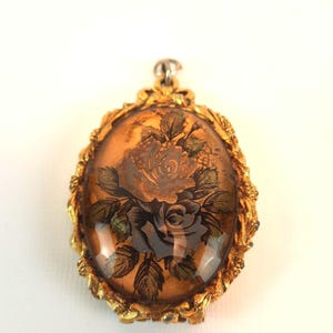 May include: An oval-shaped pendant featuring a gold-toned metal frame with intricate leaf-like detailing. The center contains a translucent amber-colored insert decorated with a dark, detailed illustration of two blooming roses and foliage.