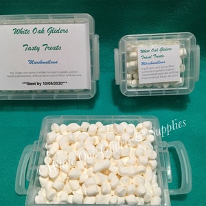 May include: Three clear plastic containers filled with white marshmallows. The containers have labels that read "White Oak Gliders" and "Tasty Treats" or "Travel Treats". The text "Best by 10/05/2020" is also visible.