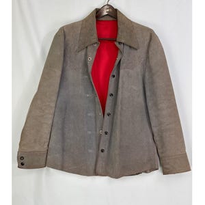 Op de afbeelding: Een vintage bruin suède jasje met een rode voering. De jas heeft een knoopsluiting aan de voorkant, een kraag en manchetten met knopen. De jas hangt aan een houten kleerhanger tegen een witte achtergrond.