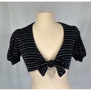 May include: A black cropped bolero cardigan featuring thin horizontal silver stripes, short puff sleeves with ribbed cuffs, and a front tie closure. The garment has a deep V-neckline and is displayed on a mannequin to show the structure and fit.