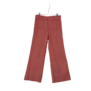 Puede incluir: Un pantalón de color coral, de pierna ancha con finas rayas verticales, colgado de una percha metálica. El pantalón tiene dos bolsillos delanteros y un cierre de botón. La tela parece ser un material ligero.