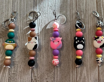 May include: Five beaded keychains with silver-tone hardware. Each keychain features a different animal charm: a penguin, a cat, a pig, a black cat, and a cow. The beads are in various colors, including brown, green, pink, purple, and black.
