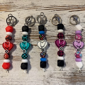 May include: Five keychains with silver rings and various bead designs. Each keychain features a heart-shaped charm with a stethoscope design. The beads are in red, black, white, teal, and purple, with patterned accents.