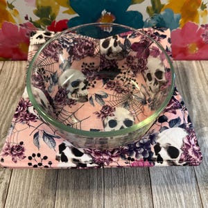 May include: A clear glass bowl sits on a square, quilted bowl cozy. The cozy features a pink background with a pattern of skulls, spiderwebs, and floral elements in shades of purple and black. The bowl cozy is designed to protect surfaces from heat.
