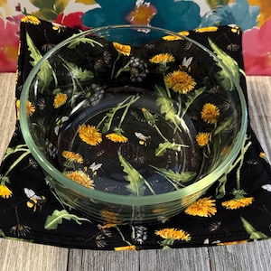May include: A clear glass bowl sits on a black bowl cozy. The bowl cozy is decorated with a pattern of yellow dandelions, green leaves, and bees. The background is a floral pattern.
