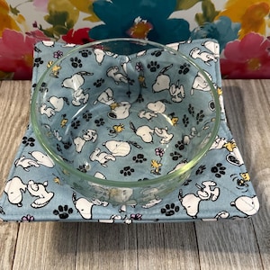 May include: A clear glass bowl sits atop a light blue fabric bowl cozy. The cozy is patterned with white Snoopy characters, black paw prints, and yellow birds. The cozy is square-shaped and designed to hold a bowl of food.