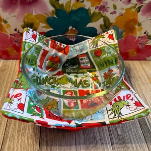 May include: A red and green fabric bowl cozy with a Grinch Christmas theme. The cozy features a variety of images from the Grinch movie, including the Grinch, Cindy Lou Who, and the Grinch's dog, Max. The cozy is perfect for keeping your bowls warm or cold.