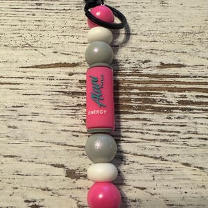 K&ouml;nnte beinhalten: Ein Schl&uuml;sselanh&auml;nger mit einem schwarzen Metallverschluss und Ring, befestigt an einer vertikalen Anordnung von Perlen. Die Perlen umfassen rosa, wei&szlig;e und schimmernde graue Kugeln sowie eine zentrale rosa zylindrische Perle mit dem Text Alani ENERGY.