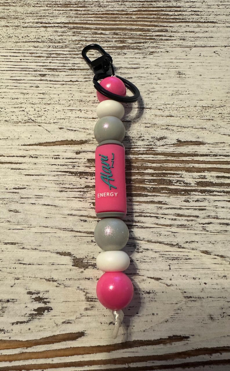 K&ouml;nnte beinhalten: Ein Schl&uuml;sselanh&auml;nger mit einem schwarzen Metallverschluss und Ring, befestigt an einer vertikalen Anordnung von Perlen. Die Perlen umfassen rosa, wei&szlig;e und schimmernde graue Kugeln sowie eine zentrale rosa zylindrische Perle mit dem Text Alani ENERGY.