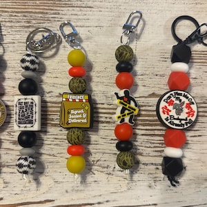 May include: A collection of beaded keychains with various charms. The keychains feature a mix of round and geometric beads in colors like red, black, white, yellow, and green. Charms include text and graphic designs related to crime scenes and evidence.