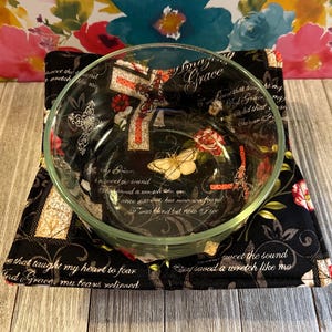 May include: A clear glass bowl sits on a black bowl cozy. The cozy features a floral and butterfly design with text. The bowl cozy is square and has red and white accents. The background is a floral pattern.