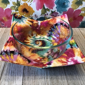May include: A clear glass bowl sits in a colorful tie-dye bowl cozy. The cozy features a vibrant pattern of pink, yellow, orange, and teal hues. The cozy is square-shaped with stitched edges, designed to hold a bowl.