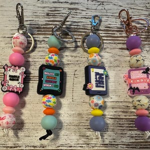 May include: Four beaded keychains featuring silicone beads in various colors and shapes, each with a central charm shaped like an e-reader. The charms display text including Kindle Girlie, Delete My Kindle History When I Die, and A Reader Lives A Thousand Lives.