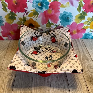 May include: A clear glass bowl sits on a red and white Mickey Mouse-themed bowl cozy. The cozy is square-shaped with a red border and a pattern of Mickey Mouse figures in black, white, and red.