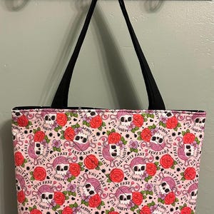 May include: A pink tote bag with a black base and handles. The bag features a repeating pattern of skulls, roses, and yarn balls. The words "Knit Fast, Die Warm" are also printed on the bag.