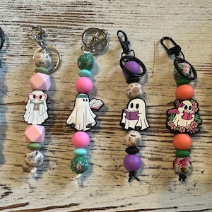 May include: Six beaded keychains featuring ghost-themed charms. Each keychain includes a metal clasp and a variety of silicone and patterned beads in colors like orange, pink, green, black, and purple, with ghost figures reading books.
