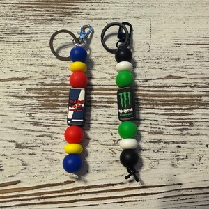 K&ouml;nnte beinhalten: Zwei Schl&uuml;sselanh&auml;nger aus Perlen mit Metallringen. Der linke Anh&auml;nger hat blaue, gelbe und rote Perlen mit einem rechteckigen Red Bull Logo-Anh&auml;nger. Der rechte Anh&auml;nger hat schwarze, wei&szlig;e und gr&uuml;ne Perlen mit einem Monster Energy Logo-Anh&auml;nger.
