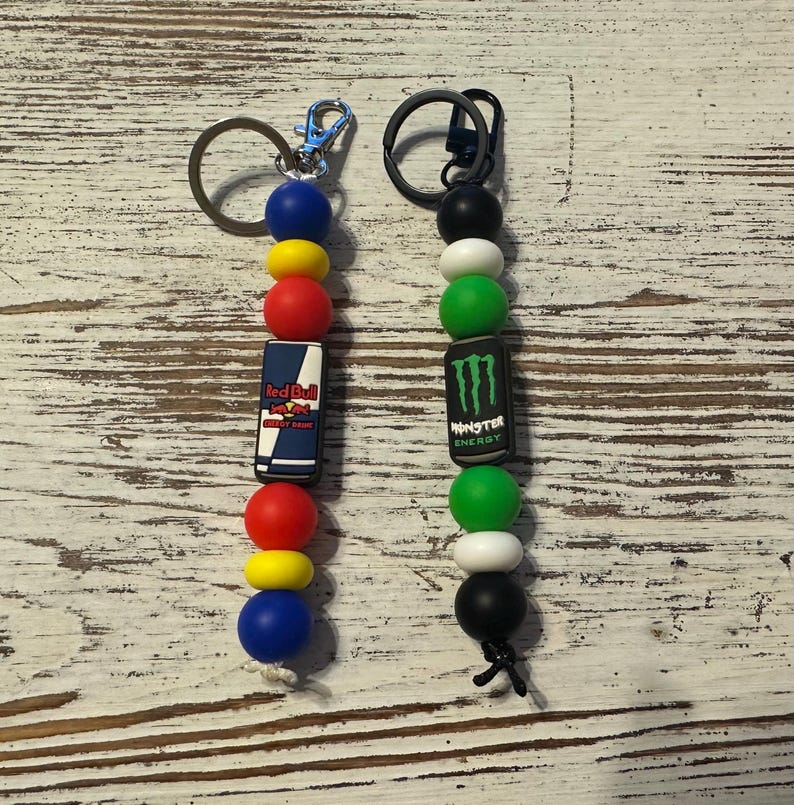 K&ouml;nnte beinhalten: Zwei Schl&uuml;sselanh&auml;nger aus Perlen mit Metallringen. Der linke Anh&auml;nger hat blaue, gelbe und rote Perlen mit einem rechteckigen Red Bull Logo-Anh&auml;nger. Der rechte Anh&auml;nger hat schwarze, wei&szlig;e und gr&uuml;ne Perlen mit einem Monster Energy Logo-Anh&auml;nger.