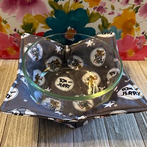 May include: A gray and white fabric bowl cozy with a repeating pattern of Tom and Jerry cartoon characters and stars. The cozy is holding a clear glass bowl.