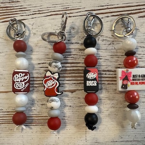 May include: Four keychains with Dr. Pepper-themed charms. Each keychain features a silver clasp, red, white, and black beads, and a charm with the Dr. Pepper logo or a related phrase. One charm reads "Just a girl obsessed with her Dr. Pepper."