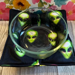 May include: A clear glass bowl resting inside a square fabric bowl cozy. The fabric features a black background with a repeating pattern of green alien heads with large black eyes. The bowl cozy has stitched edges and a quilted texture throughout.