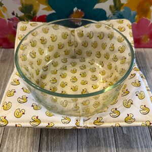 May include: A clear glass bowl sits on a square, quilted bowl cozy. The cozy is white with a repeating pattern of yellow rubber duckies. The interior of the bowl also features the same ducky pattern. The background is a floral pattern.