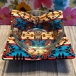 May include: A square, quilted hot pad with a Native American-inspired geometric pattern in shades of red, blue, black, and beige. A clear glass bowl sits on top of the hot pad.