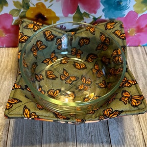 May include: A green fabric bowl cozy with a butterfly pattern. The cozy is filled with a clear glass bowl.