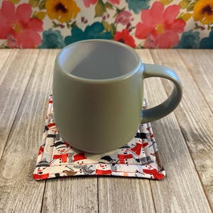 May include: A gray ceramic mug with a handle sits on a square coaster with a white background and a pattern of snowmen wearing red scarves and hats.