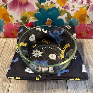 May include: A clear glass bowl sits on a black, square, heat-resistant pad. The pad features a repeating pattern of a blue cartoon character, white speech bubbles, and yellow text. The bowl's interior also has a similar design.