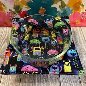 May include: A black bowl cozy with a colorful monster print. The cozy is made of fabric and has a quilted design. The bowl is clear glass and is sitting inside the cozy.