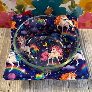 May include: A clear glass bowl rests on a navy blue, square, padded bowl cozy. The bowl and cozy feature a whimsical design of unicorns, rainbows, planets, and stars. The cozy is designed to protect surfaces from hot bowls.
