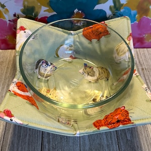 May include: A green fabric bowl cozy with a guinea pig print. The cozy has a glass bowl inside.