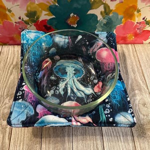 May include: A glass bowl with a black background and a colorful jellyfish pattern sits on a black and white fabric bowl cozy.