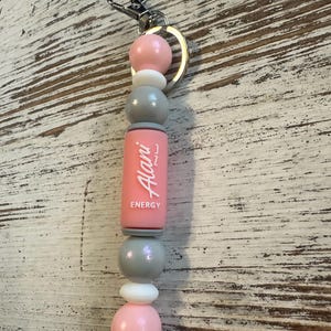 K&ouml;nnte beinhalten: Ein Schl&uuml;sselanh&auml;nger mit einer Reihe von runden und zylindrischen Perlen in Rosa, Grau und Wei&szlig;. Eine zentrale rosa zylindrische Perle zeigt den wei&szlig;en Schriftzug Alani ENERGY. Die Perlen sind aufgereiht und an einem silbernen Metallring befestigt.