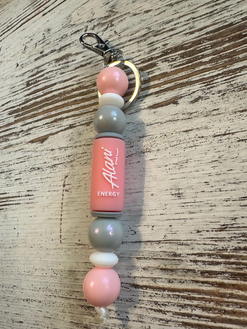 K&ouml;nnte beinhalten: Ein Schl&uuml;sselanh&auml;nger mit einer Reihe von runden und zylindrischen Perlen in Rosa, Grau und Wei&szlig;. Eine zentrale rosa zylindrische Perle zeigt den wei&szlig;en Schriftzug Alani ENERGY. Die Perlen sind aufgereiht und an einem silbernen Metallring befestigt.