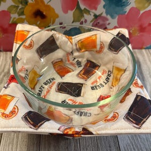 May include: A clear glass bowl rests on a square, padded, cream-colored fabric. The fabric is printed with various beer glasses and the words "Craft Beer Served Here." The background is a floral pattern with pink, yellow, and blue flowers.