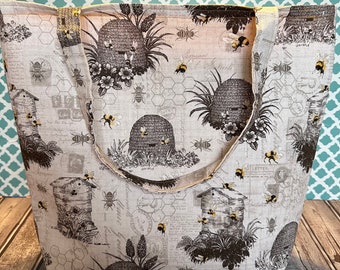 May include: A beige tote bag with a white background and a pattern of brown beehives, bees, and flowers. The bag has two brown straps.