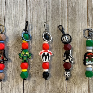 May include: Five beaded keychains with various charms. The charms include a wolf, a creature, a bat, a bigfoot, and a mountain scene with the text "WE BELIEVE". The beads are red, green, brown, and black.