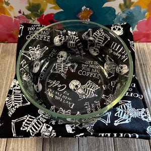 May include: A clear glass bowl sits atop a black bowl cozy. The cozy features a white skeleton and text design. The text reads "Life is too short for bad coffee" and "But first coffee". The bowl is filled with the same text and skeleton design.
