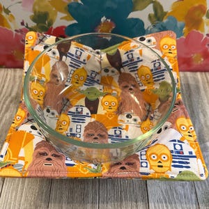 May include: A clear glass bowl sits on a square, padded bowl cozy. The cozy features a repeating pattern of Star Wars characters in shades of orange, white, and brown. The background is a floral pattern.