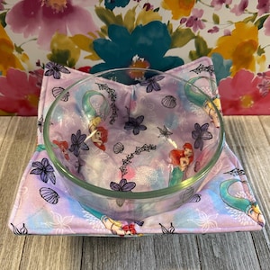 May include: A clear glass bowl rests on a lavender bowl cozy featuring a mermaid and floral design. The cozy has a quilted pattern with images of seashells and flowers. The background is a floral pattern.