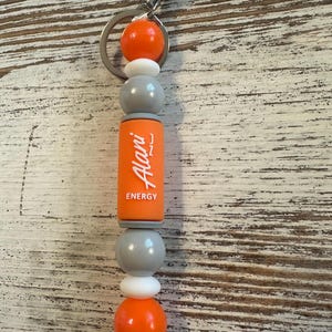 K&ouml;nnte beinhalten: Ein Schl&uuml;sselanh&auml;nger mit einem silbernen Karabinerhaken und Ring, an dem eine Reihe von Perlen befestigt ist. Die Perlen umfassen zwei gro&szlig;e orangefarbene Kugeln, zwei kleine wei&szlig;e Scheiben, zwei graue Kugeln und einen orangefarbenen Zylinder mit Alani Energy Text.