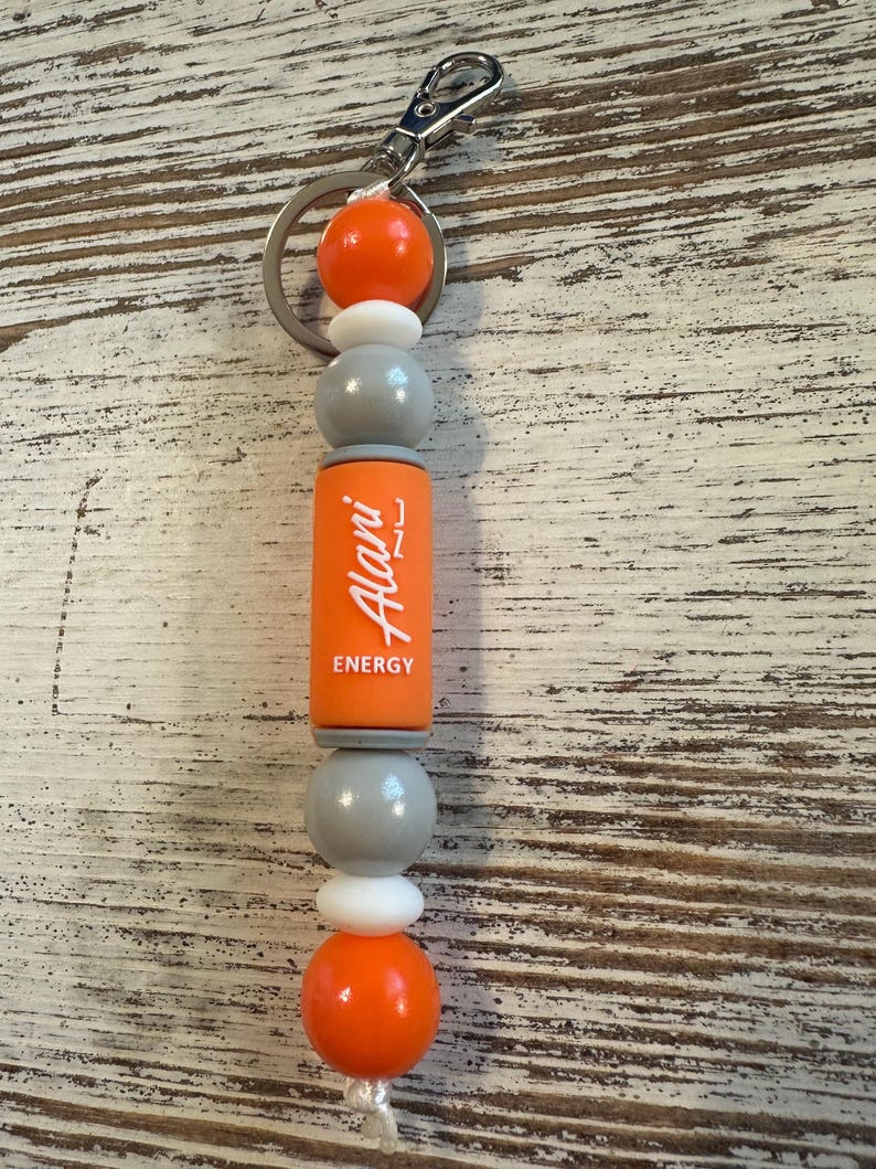 K&ouml;nnte beinhalten: Ein Schl&uuml;sselanh&auml;nger mit einem silbernen Karabinerhaken und Ring, an dem eine Reihe von Perlen befestigt ist. Die Perlen umfassen zwei gro&szlig;e orangefarbene Kugeln, zwei kleine wei&szlig;e Scheiben, zwei graue Kugeln und einen orangefarbenen Zylinder mit Alani Energy Text.