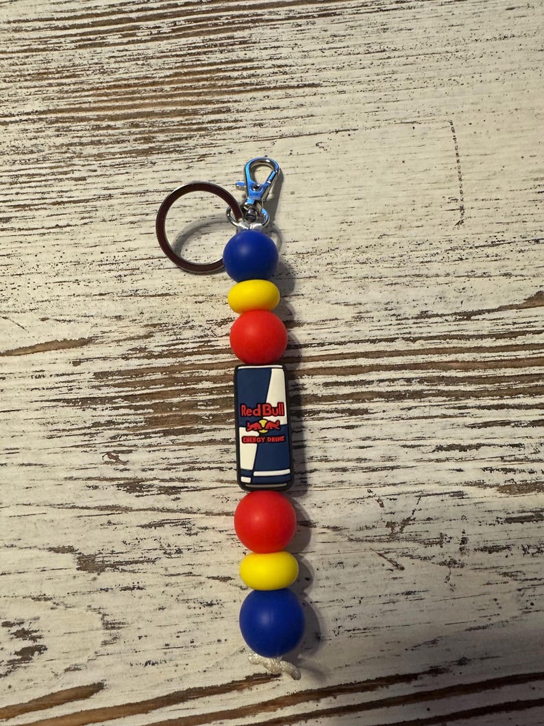 K&ouml;nnte beinhalten: Ein Schl&uuml;sselanh&auml;nger mit einem zentralen rechteckigen Anh&auml;nger mit dem Red Bull Energy Drink Logo, flankiert von abwechselnd blauen, gelben und roten kugelf&ouml;rmigen und scheibenf&ouml;rmigen Perlen. Der Anh&auml;nger ist an einem silberfarbenen Karabinerhaken befestigt.