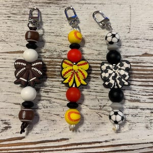 May include: Three beaded keychains with sports themes. One features a football, a brown bow, and white and black beads. Another has a softball, a yellow bow, and red and black beads. The third has a soccer ball, a black and white bow, and black and gray beads.